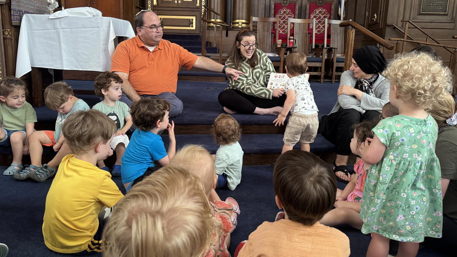 Children gathered for story time and group learning in a synagogue sanctuary with rabbis.