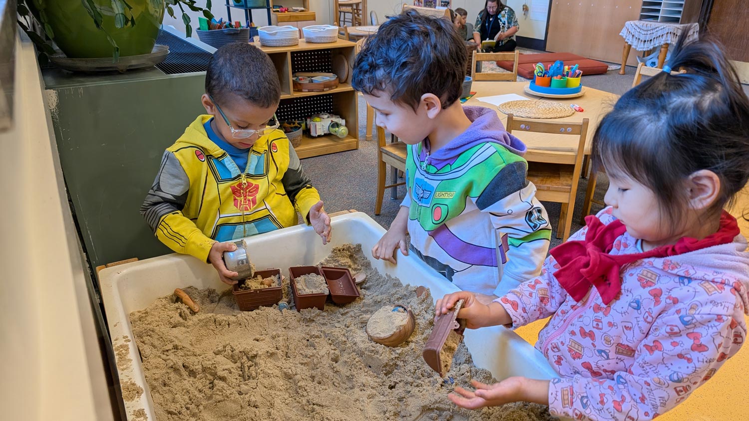Children participating in sensory play at a classroom sand table, exploring and building with scoops, containers, and molds.