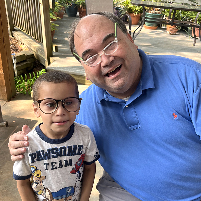 Adult and child wearing oversized eyeglasses pose together in an outdoor garden setting.