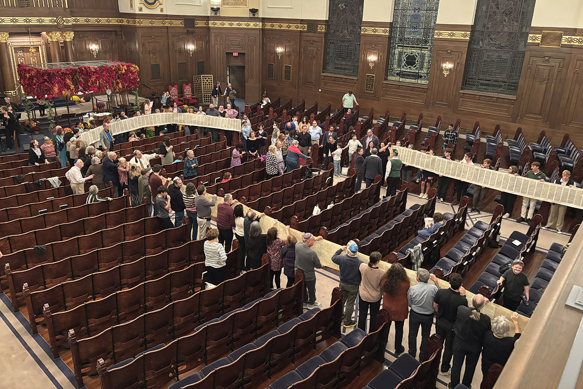 Congregants of all ages hold an unrolled Torah scroll around the sanctuary during a Simchat Torah service, creating a continuous display of the entire scroll.