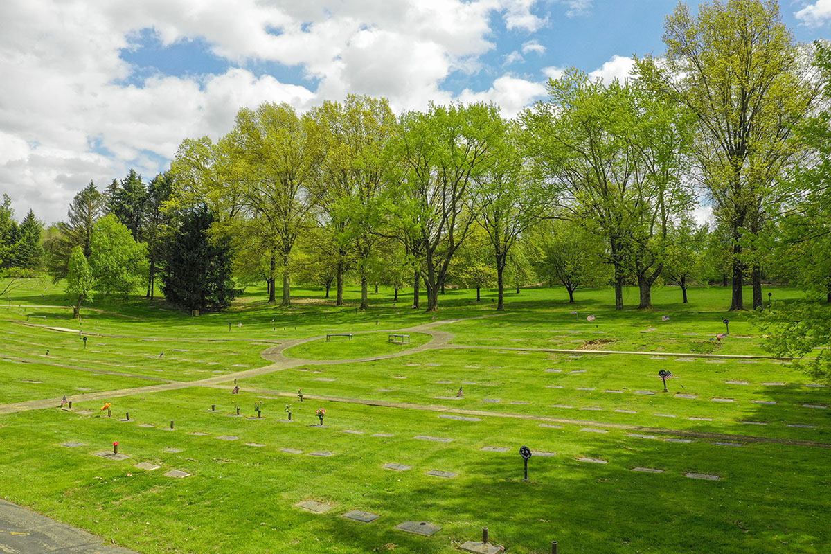 Memorial Park synagogue cemetery with flat memorial markers, green lawns, winding pathways, and mature trees under a partly cloudy sky.