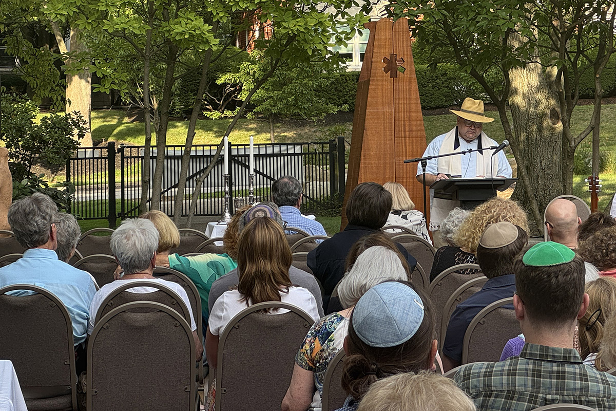 Rabbi Fellman wearing a straw hat leading an outdoor Shabbat service with congregants seated with their backs to the camera.