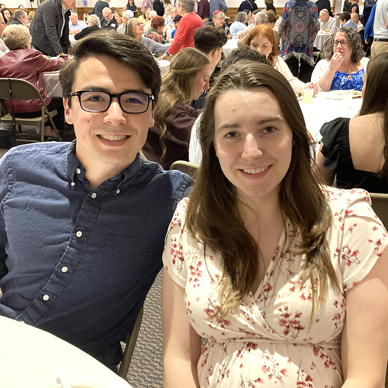 Young adult couple seated together at a Shabbat dinner, with community members gathered around tables in the background.