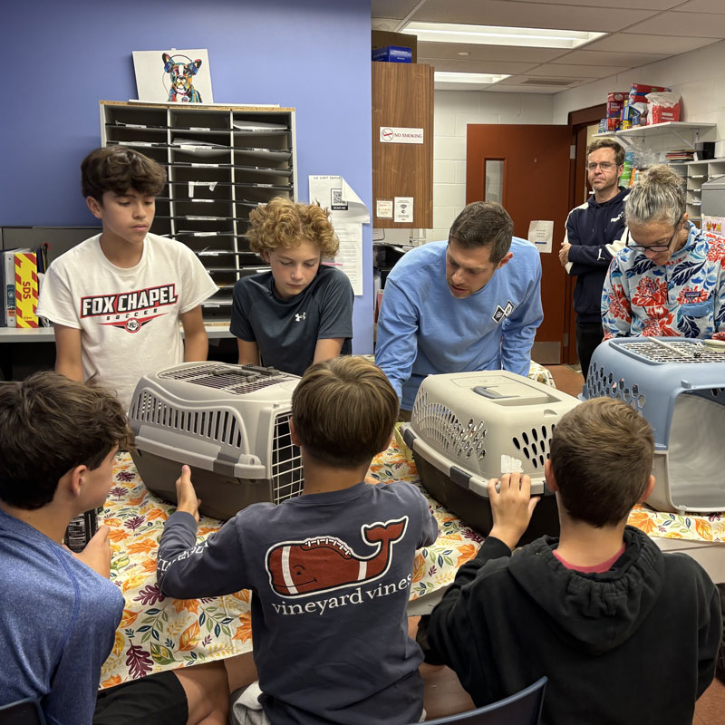 Teens and adults gather around a table lined with pet carriers at an animal shelter during a social action program.